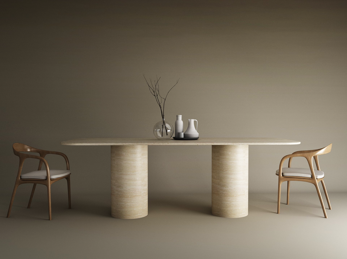 The TRAVERTABLE Rectangular Travertine Dining Table with Cylindrical Base sits between two wooden chairs with white cushions. On the natural stone surface are a clear vase with branches and three ceramic vessels, set against a plain brown wall.