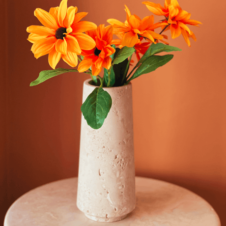 A beige ceramic vase holds bright orange flowers with green leaves, placed on a round marble table against a matching orange background.