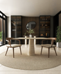 A modern dining room featuring the TRAVERTABLE Square Travertine Dining Table with Conical Pedestal Base, two wooden chairs, a circular rug, built-in shelves with decor, a round mirror on a wood-paneled wall, large windows, and green plants.