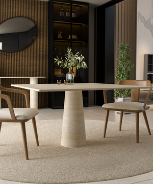 A modern dining area featuring the TRAVERTABLE Square Travertine Dining Table with a conical pedestal base, two wooden cushioned chairs, a vase with branches, a circular rug, a plant, and shelves with bottles and glasses in the background.