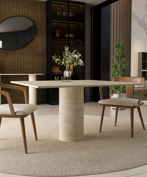 A modern dining room showcases the TRAVERTABLE Square Travertine Dining Table with Cylindrical Center Base, two cushioned wooden chairs, a vase with green branches, carpeted floor, glassware shelves, wall mirror, and a tall plant.