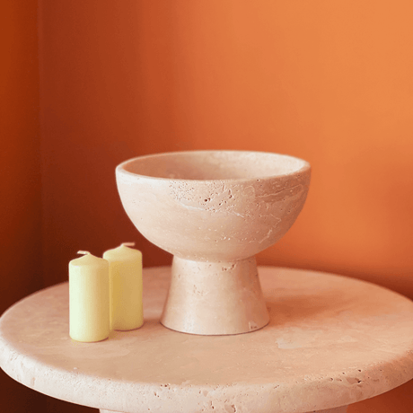 The Sleek Modern Travertine Marble Bowl by Travertable sits on a matching light beige circular table, accompanied by two unlit, pale yellow pillar candles, all set against a warm orange wall.