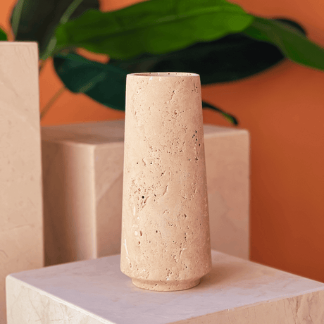 The Classic Beige Travertine Vase by Travertable sits tall on a marble platform, against an orange background with large green leaves.