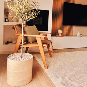 A modern living room includes a wooden armchair with green and brown cushions next to a large potted plant, complementing the Golden Sunrise Travertine Vase by Travertable. Wooden paneling and a white TV console with a flat-screen TV accentuate the scene, while a textured rug adorns the floor.
