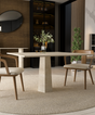A modern dining area showcases the TRAVERTABLE Square Travertine Dining Table with Rockcone Center Base, two cushioned wooden chairs, a greenery-filled vase, and a round beige rug. A dark shelf, round mirror, and potted plant accent the background.