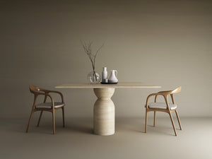 The Square Travertine Dining Table with Sculptural Round Center Base by TRAVERTABLE, featuring an organic stone form, is styled with vases and a pitcher. Two wooden chairs with white cushions flank this customizable table against a beige backdrop.