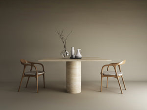 A minimalist dining setup featuring TRAVERTABLEs Square Travertine Dining Table with Cylindrical Center Base in pure stone, paired with two wooden chairs with white cushions and vases, set against a plain beige background.