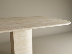 The TRAVERTABLE Rectangular Travertine Dining Table features a natural stone top with rounded corners and a thick sloped pedestal base, all crafted from solid travertine, standing out against a plain neutral background. Customization available.