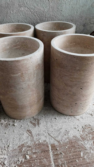 Four Golden Sunrise Travertine Vases by Travertable, featuring a smooth, light brown surface and subtle texture, stand on a dusty surface with a gray, uneven background.