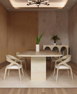 A modern dining room features the TRAVERTABLE Rectangular Travertine Dining Table with a travertine stone base, four light wood chairs with white cushions, a vase of green leaves, and a sideboard with vases against textured beige walls.