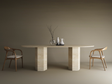 A minimal dining setup features the TRAVERTABLE Oval Travertine Dining Table with a hexagonal stone base, two wooden chairs with white cushions, three ceramic vessels, and a glass vase with branches, all set against a plain brown background.