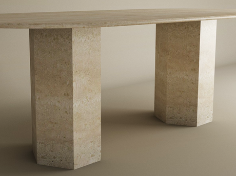 The TRAVERTABLE Oval Travertine Dining Table features a natural stone tabletop and a hexagonal organic stone pedestal base in beige with a textured finish. The background is plain and neutral. Customization available.