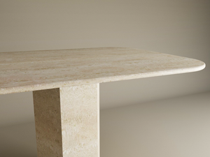 A close-up of the TRAVERTABLE Rectangular Travertine Dining Table, featuring a minimalist light-colored top, rounded edges, and an organic hexagonal stone pedestal base, set against a neutral beige background.