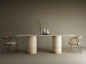 The TRAVERTABLE Rectangular Travertine Dining Table with Cylindrical Base sits between two wooden chairs with white cushions. On the natural stone surface are a clear vase with branches and three ceramic vessels, set against a plain brown wall.